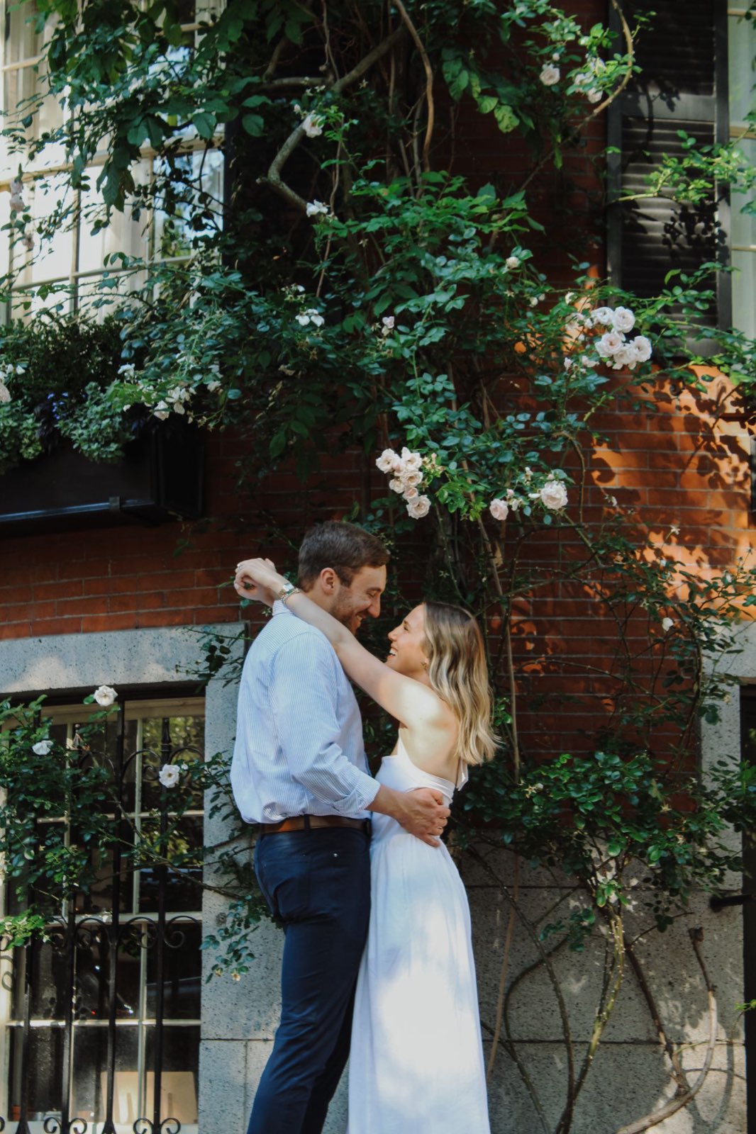 Engaged couple embracing beneath climbing roses on a red brick townhouse in Beacon Hill, Boston
