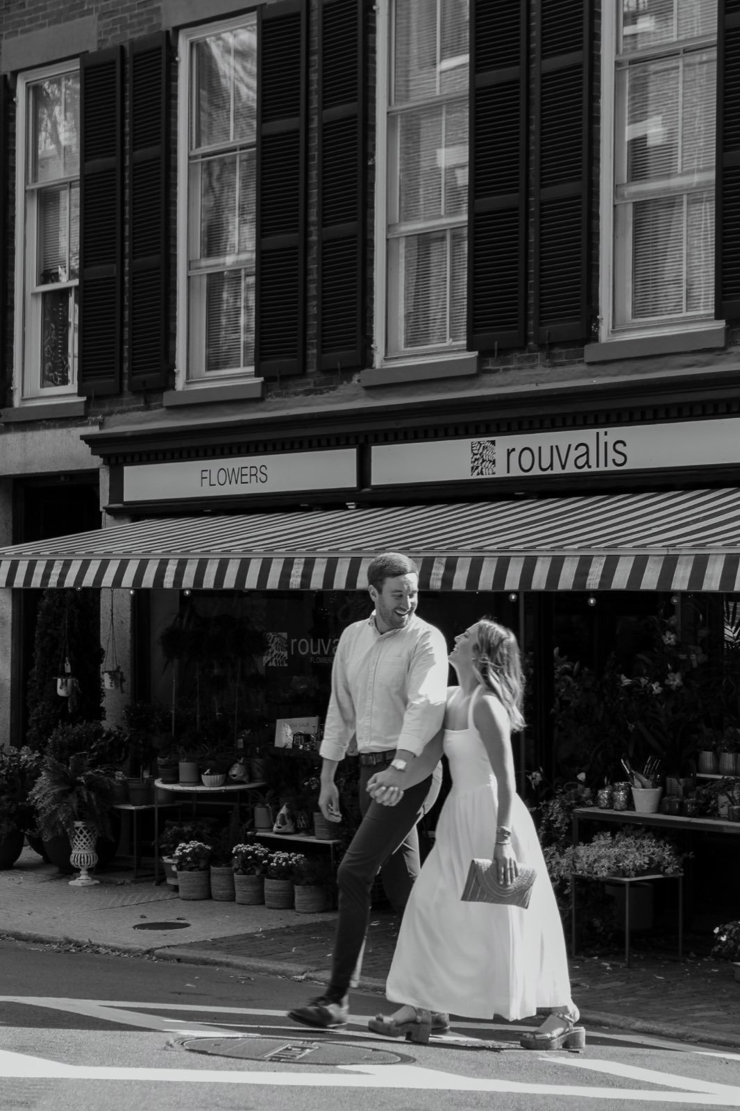 Black-and-white photo of a laughing couple walking hand in hand past a flower shop's striped awning in Beacon Hill, Boston