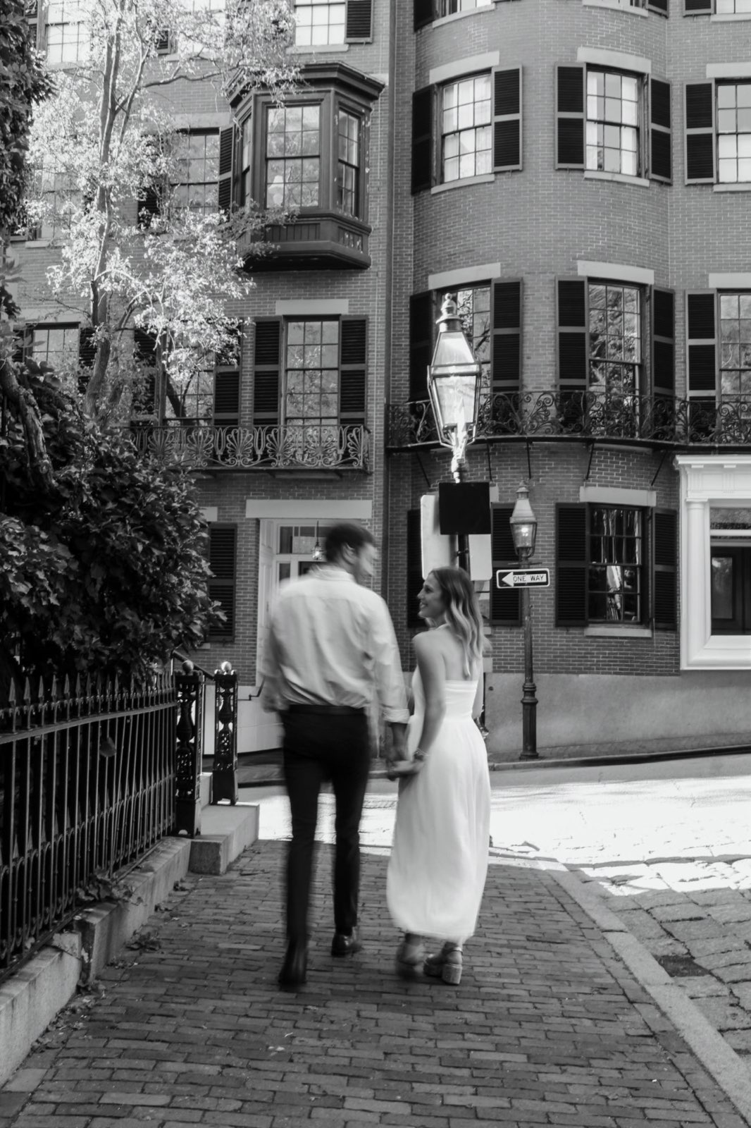 Black-and-white photo of a couple strolling down a brick sidewalk past gas lamps and iron balconies in Beacon Hill, Boston