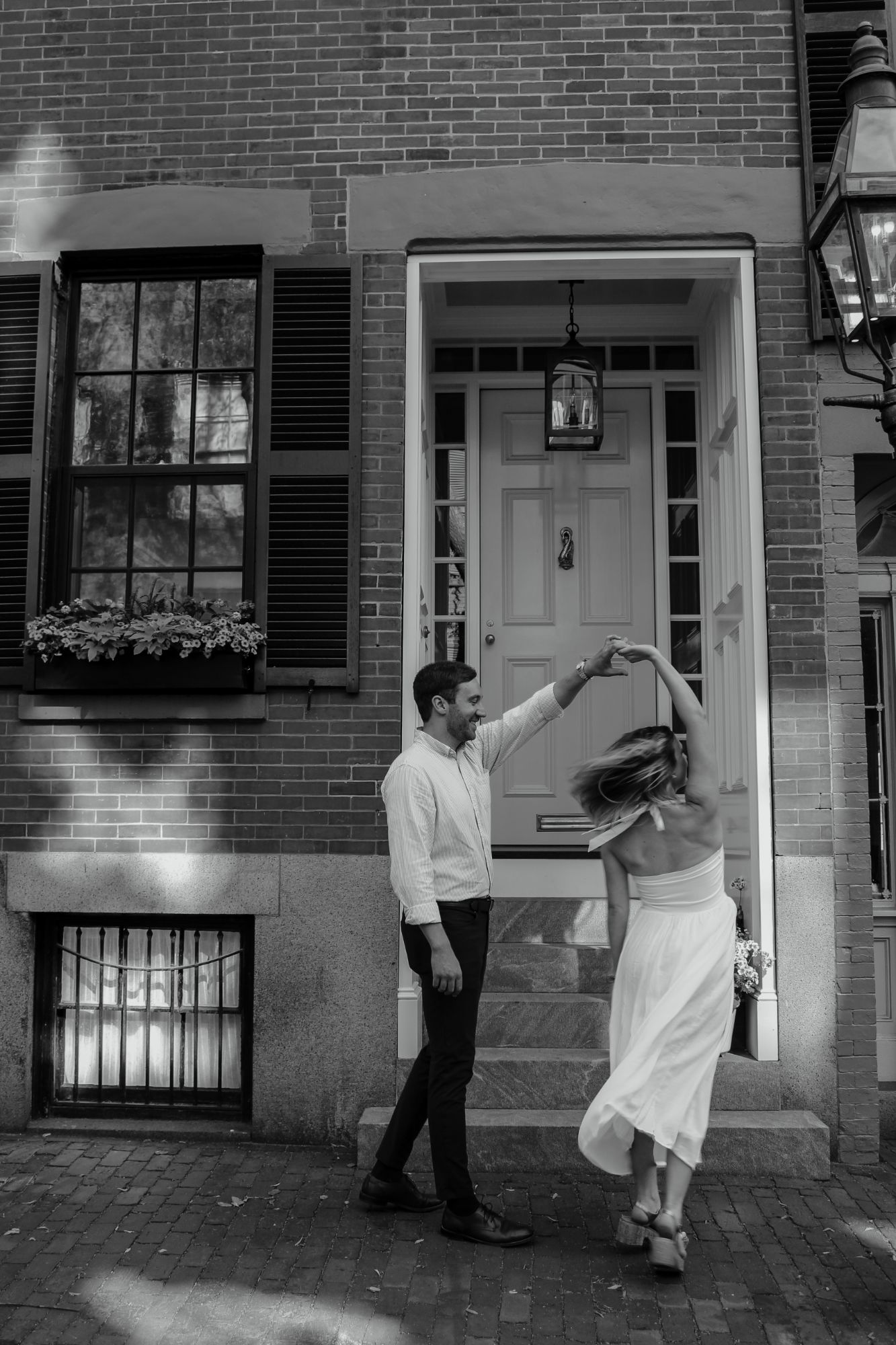 Black-and-white engagement photo of a couple twirling on a Beacon Hill sidewalk in Boston