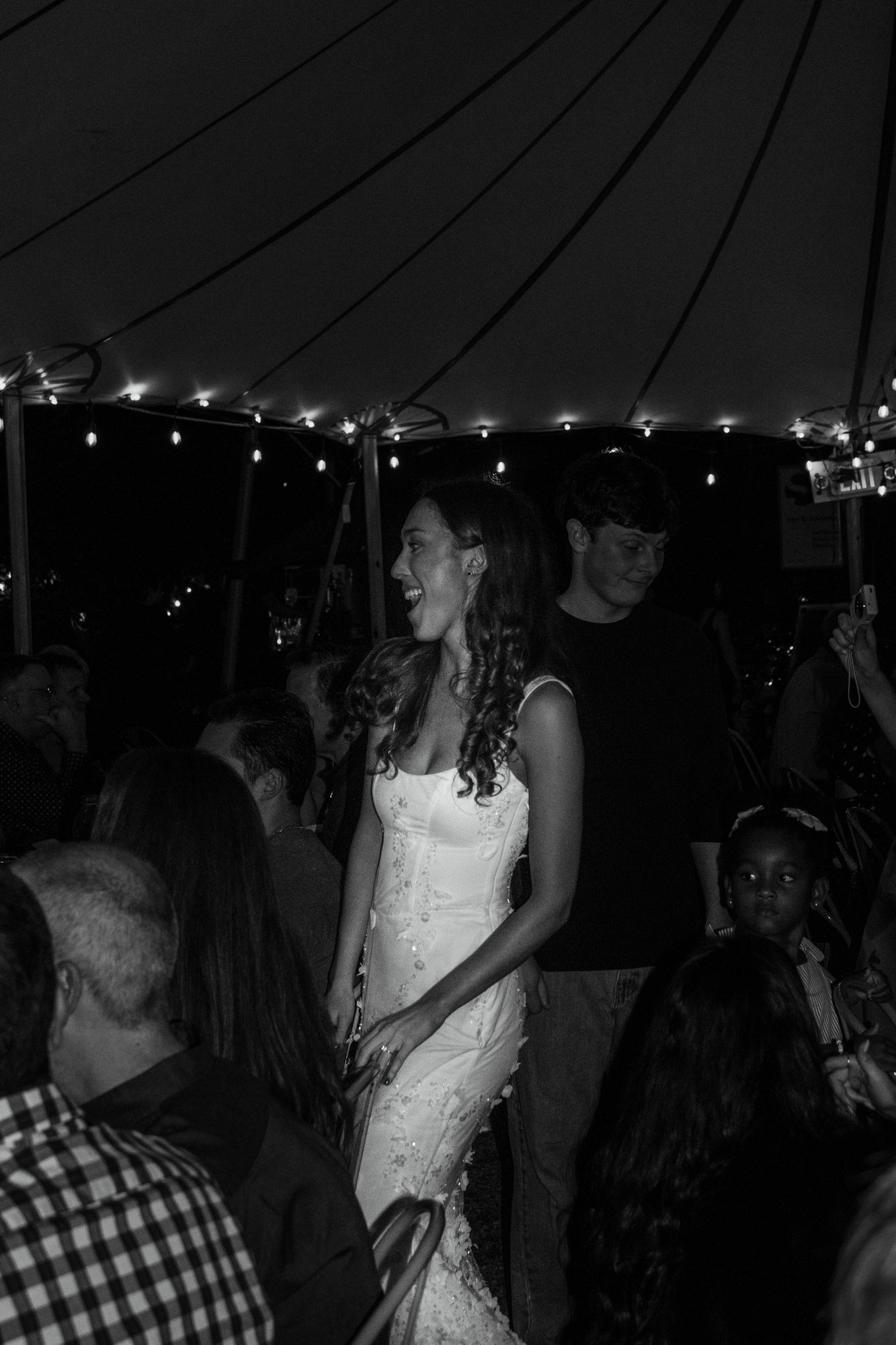 Black-and-white photo of a woman in a white dress laughing as she walks between tables under the tent at Otto Pizza, Chester