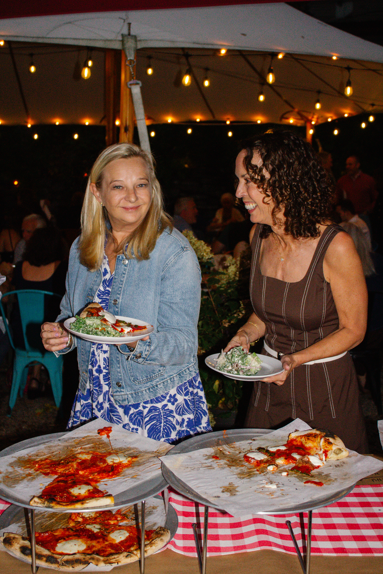 Two guests smiling as they fill their plates with pizza and salad under string lights at Otto Pizza, Chester