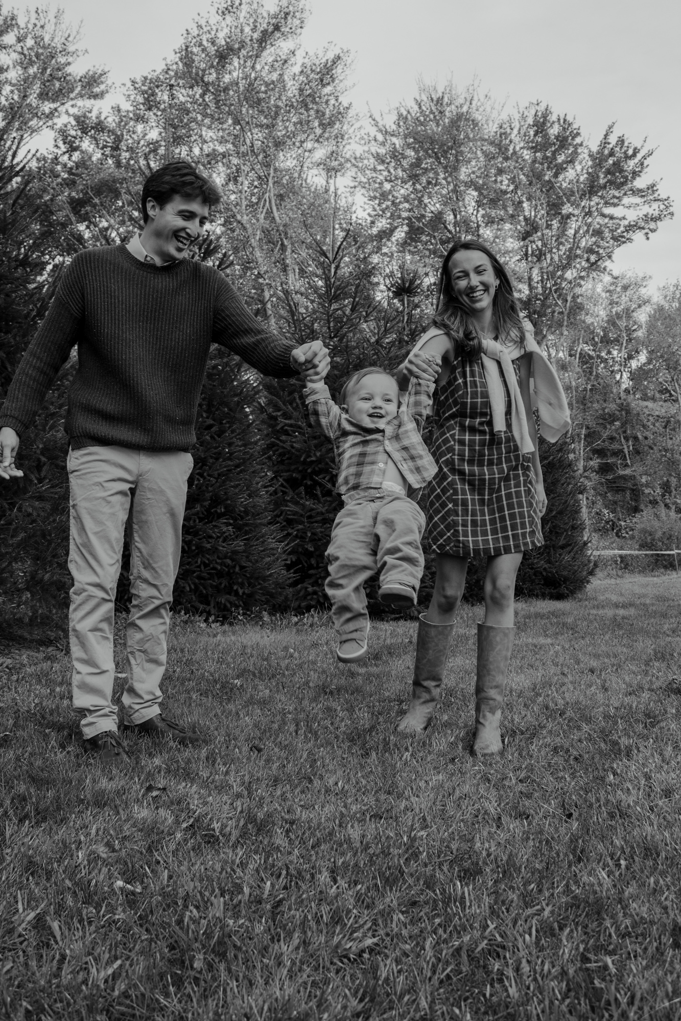 Black-and-white photo of parents swinging their toddler son between them in front of evergreens at Silverman's Farm, Easton